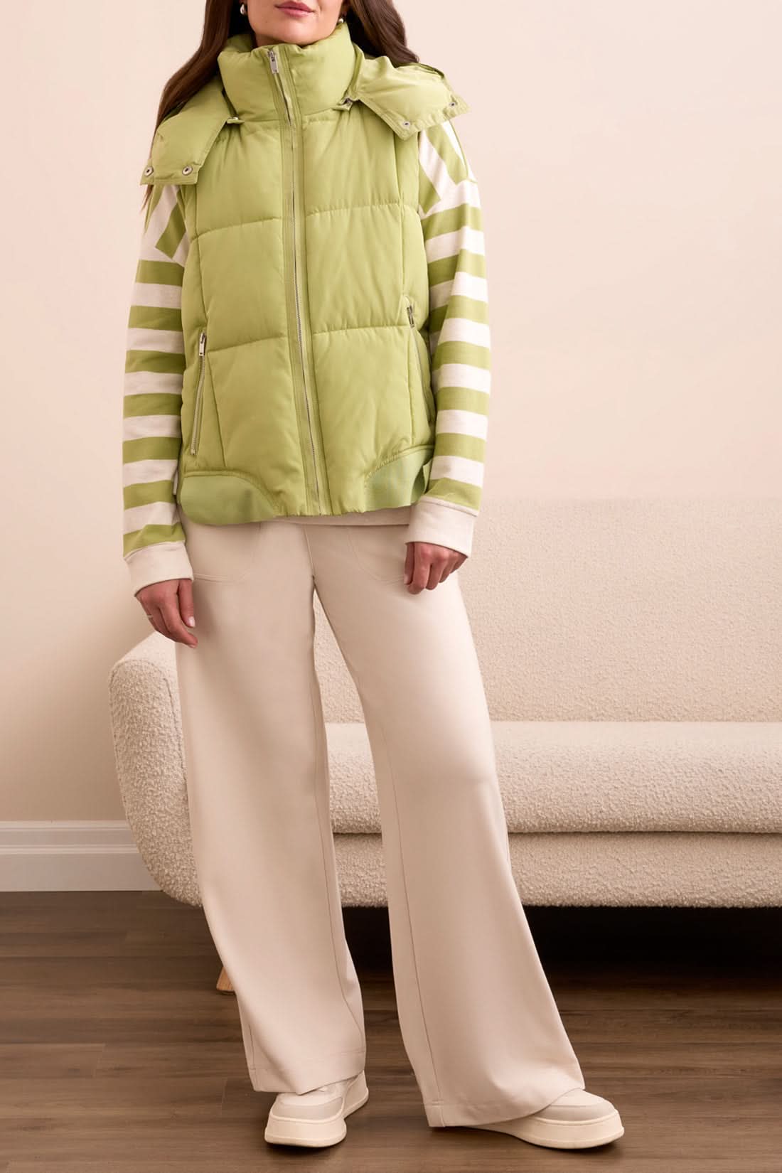 A woman stands with her hands in her pockets, looking down, wearing the Tribal QUILTED VEST in wasabi over a green and white striped sweatshirt, set against a patterned tile background.