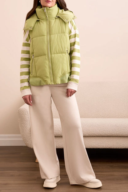 A woman stands with her hands in her pockets, looking down, wearing the Tribal QUILTED VEST in wasabi over a green and white striped sweatshirt, set against a patterned tile background.