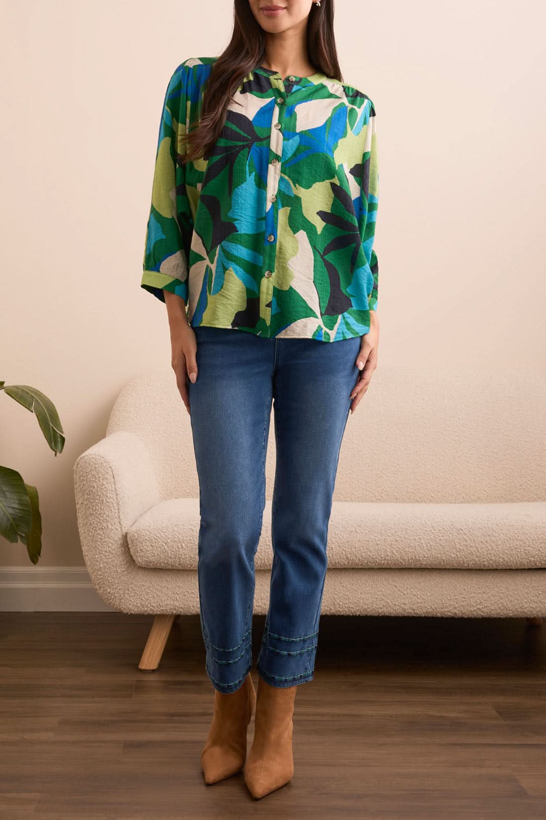A woman stands indoors in front of a cream sofa and plant, wearing the Tribal Flowy Dolman Sleeve Blouse with blue jeans and tan ankle boots.