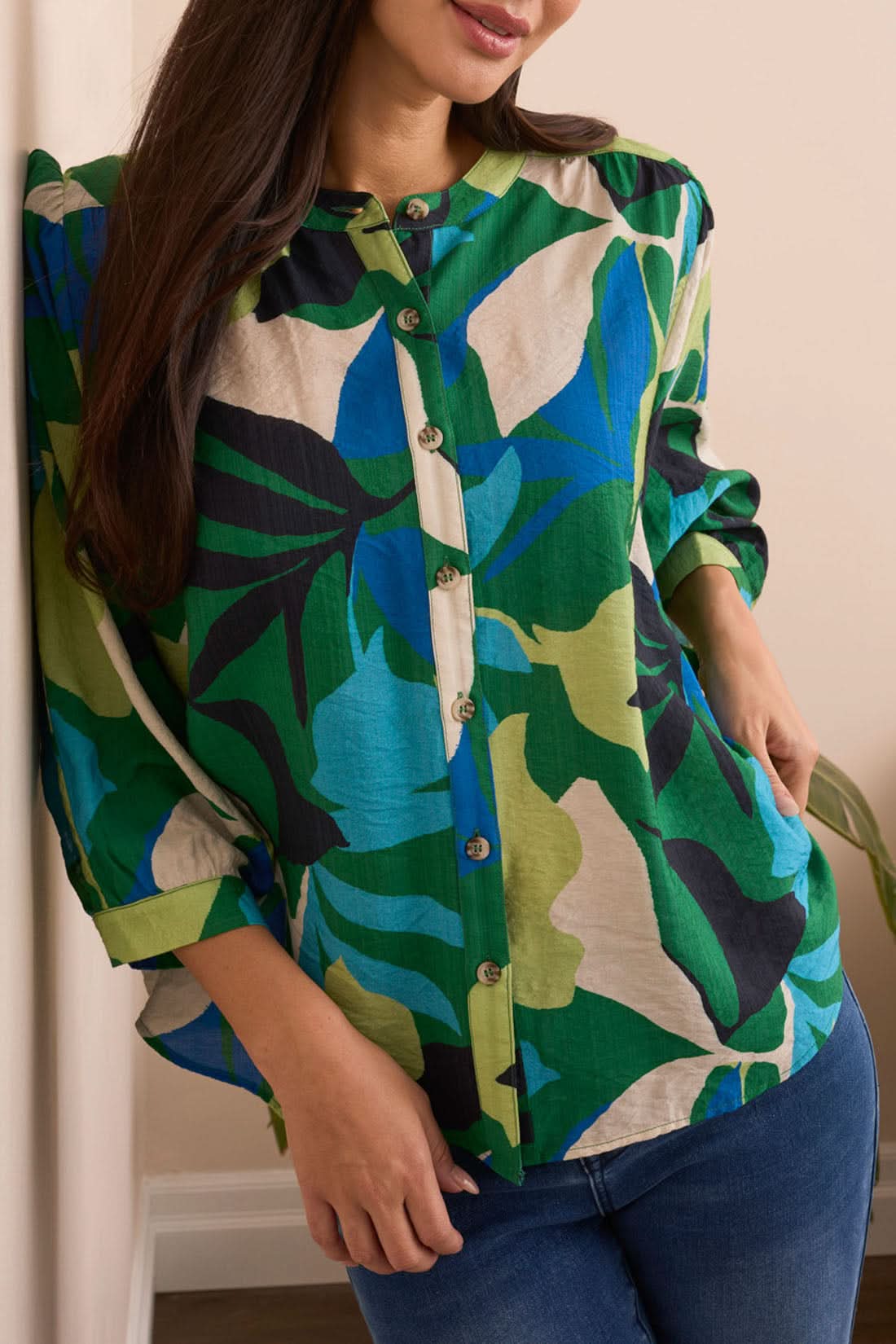 A woman stands indoors in front of a cream sofa and plant, wearing the Tribal Flowy Dolman Sleeve Blouse with blue jeans and tan ankle boots.