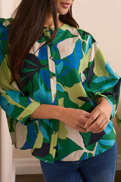 A woman stands indoors in front of a cream sofa and plant, wearing the Tribal Flowy Dolman Sleeve Blouse with blue jeans and tan ankle boots.