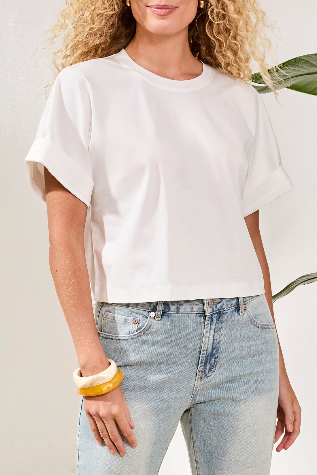 A woman stands indoors on a woven rug near a white bench and green plant, wearing the Tribal Mercerized Cotton Crew Neck T-Shirt in white, light blue straight-leg jeans, and tan heeled sandals.