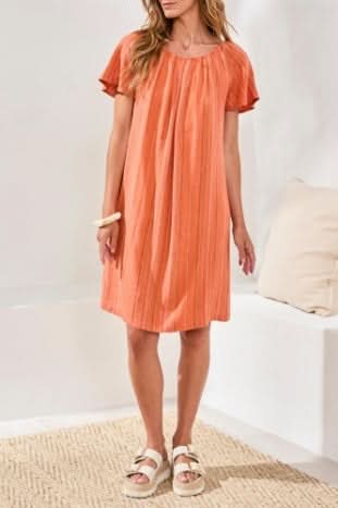 A woman wearing the Tribal FLUTTER SLV DRESS in guava blush and white sandals stands indoors on a woven rug near a white sofa with cushions.
