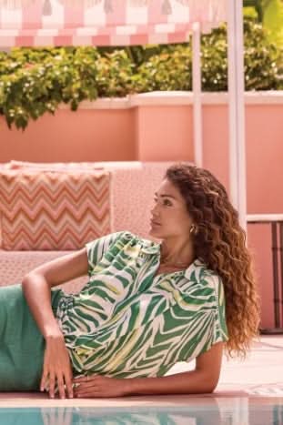 A woman with curly hair relaxes by the pool on a pink patterned cushion, wearing the Tribal Tie-Front Short-Sleeve Shirt in grass green, surrounded by greenery.