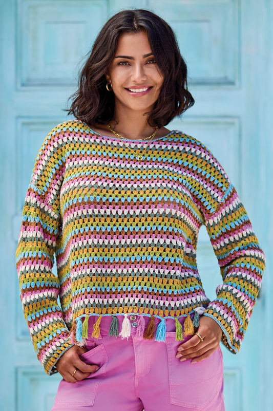 A woman smiles at the camera in front of a light blue door, wearing the Tribal Crochet Bell Sleeve Top with tassels and pink pants.