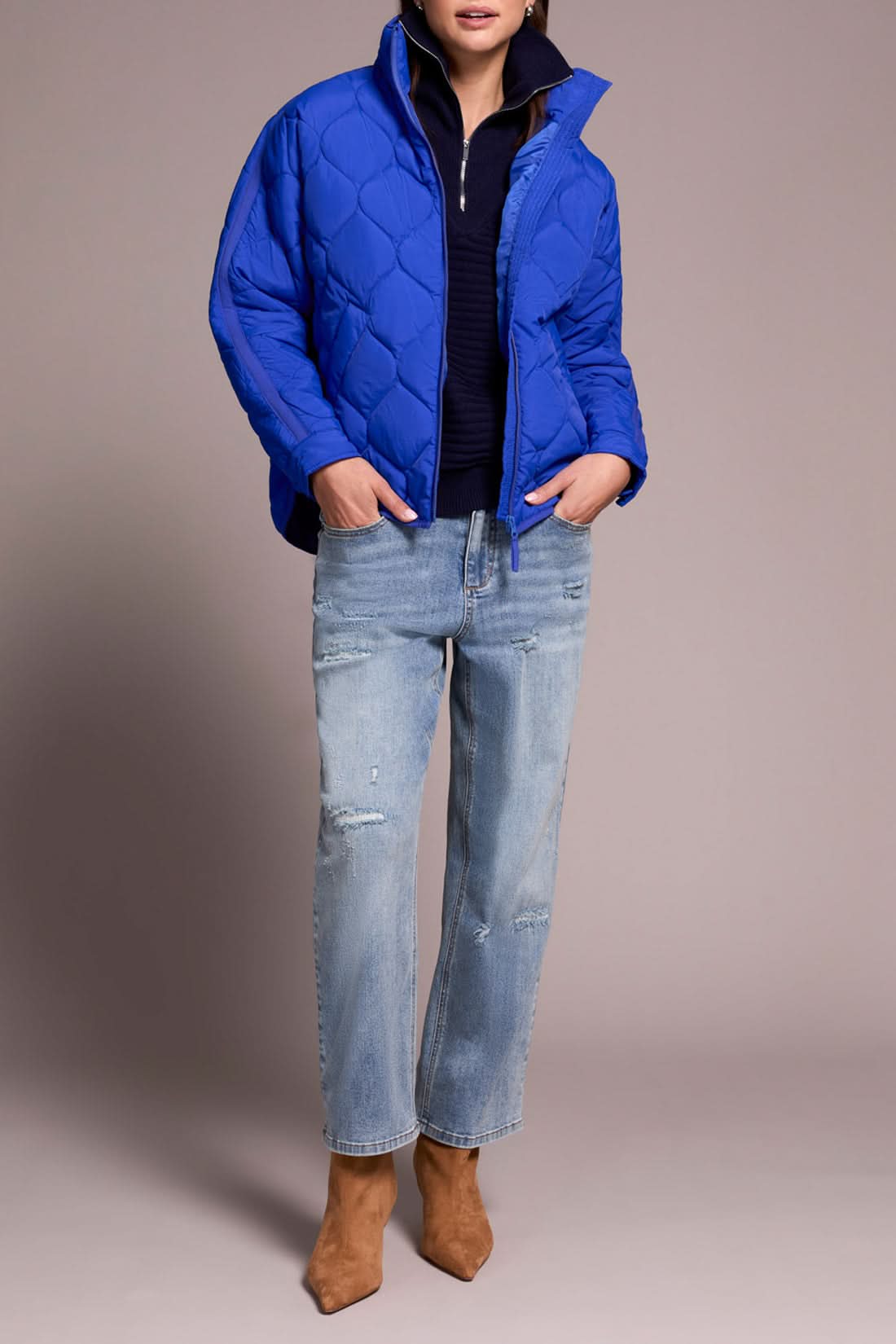 A person in the Tribal ZIPPED FRT QUILTED JACKET in lapis blue, a navy sweater, light blue distressed jeans, and tan heeled boots stands against a plain background.