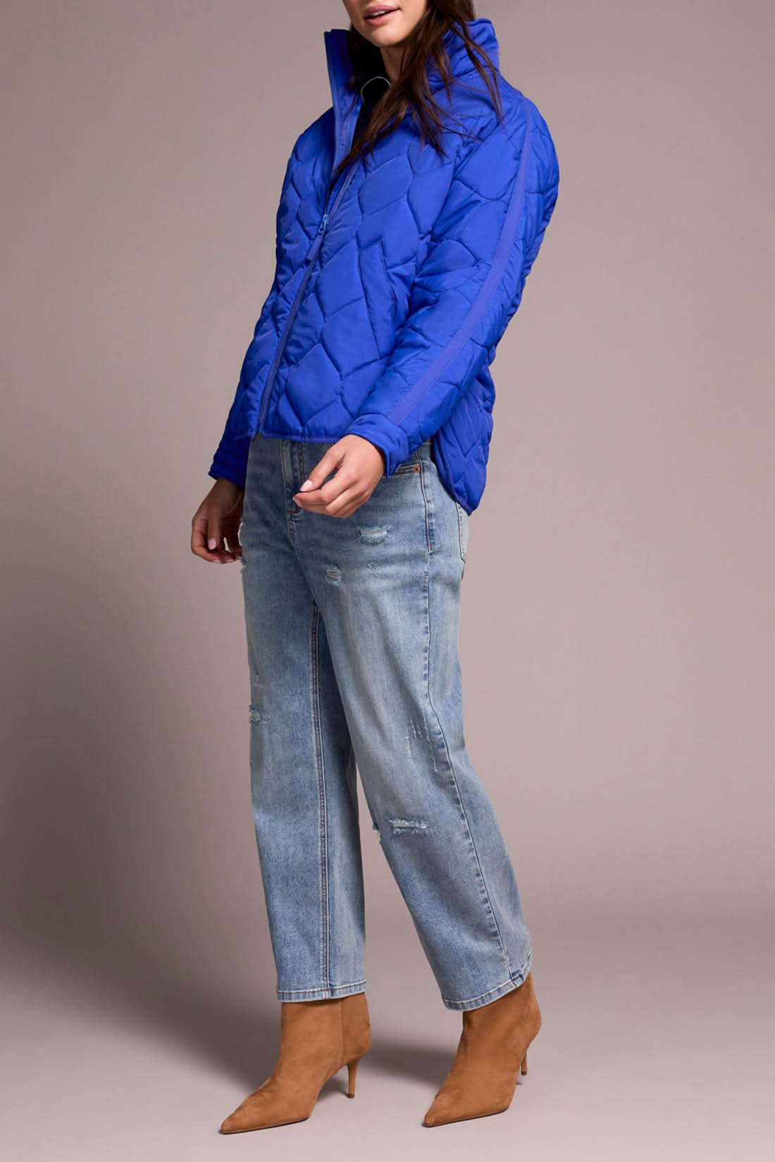 A person in the Tribal ZIPPED FRT QUILTED JACKET in lapis blue, a navy sweater, light blue distressed jeans, and tan heeled boots stands against a plain background.