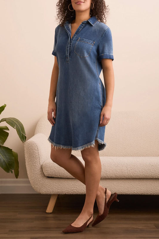 Indoors, a person wears the Tribal POP OVER SHIRT DRESS W/ POCKETS—crafted in breathable fabric with a frayed hem and front pocket. Brown shoes, hoop earrings, a plant, and a beige sofa complete the scene.