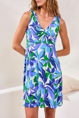 A woman stands indoors on a woven rug wearing the Tribal KNIT DRESS, a sleeveless, knee-length piece with a royal azure, green, and white abstract leaf print and flattering silhouette, paired with silver strappy sandals.