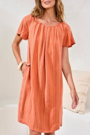 A woman wearing the Tribal FLUTTER SLV DRESS in guava blush and white sandals stands indoors on a woven rug near a white sofa with cushions.