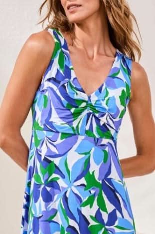A woman stands indoors on a woven rug wearing the Tribal KNIT DRESS, a sleeveless, knee-length piece with a royal azure, green, and white abstract leaf print and flattering silhouette, paired with silver strappy sandals.