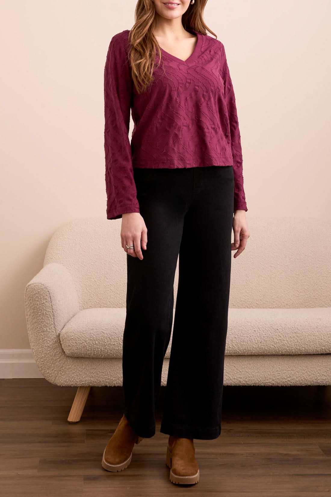 A woman stands indoors wearing the Tribal V NECK TOP in wine, paired with black wide-leg pants and brown platform shoes, with a beige couch in the background.