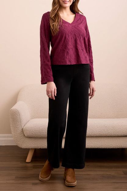 A woman stands indoors wearing the Tribal V NECK TOP in wine, paired with black wide-leg pants and brown platform shoes, with a beige couch in the background.