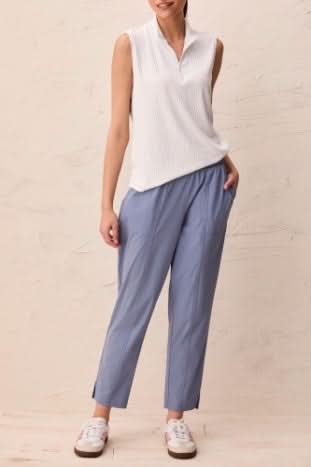 A woman stands against a neutral textured background wearing a sleeveless white blouse, Tribal PULL-ON TECHNICAL CAPRIS in light blue, and white sneakers.