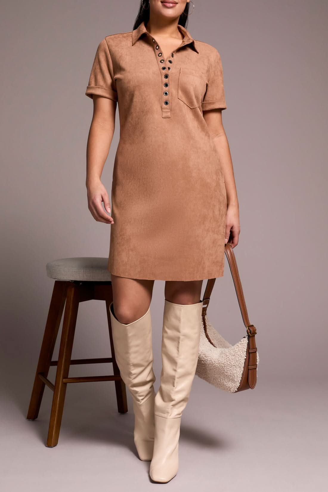 A person in the Tribal DRESS W/ FRT EYELET, paired with cream knee-high boots and a textured beige handbag, stands beside a wooden stool.
