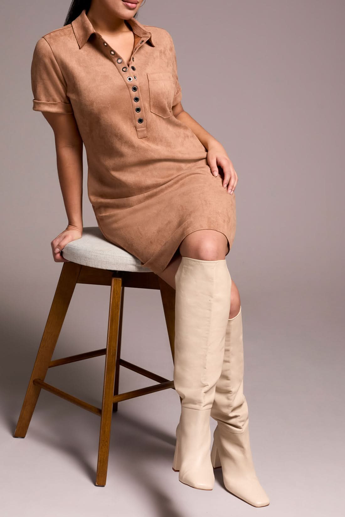 A person in the Tribal DRESS W/ FRT EYELET, paired with cream knee-high boots and a textured beige handbag, stands beside a wooden stool.