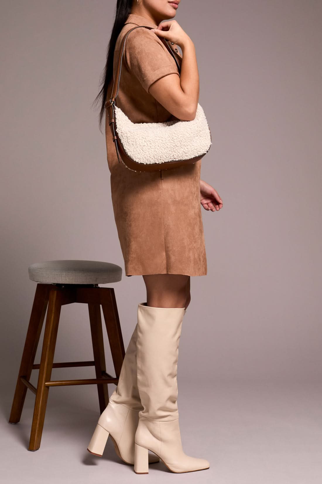 A person in the Tribal DRESS W/ FRT EYELET, paired with cream knee-high boots and a textured beige handbag, stands beside a wooden stool.