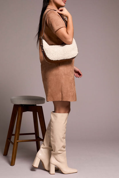 A person in the Tribal DRESS W/ FRT EYELET, paired with cream knee-high boots and a textured beige handbag, stands beside a wooden stool.