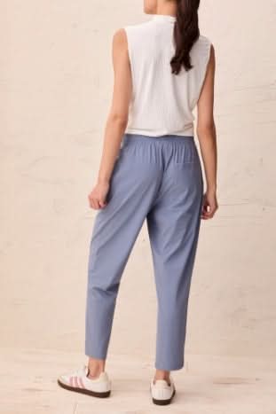 A woman stands against a neutral textured background wearing a sleeveless white blouse, Tribal PULL-ON TECHNICAL CAPRIS in light blue, and white sneakers.