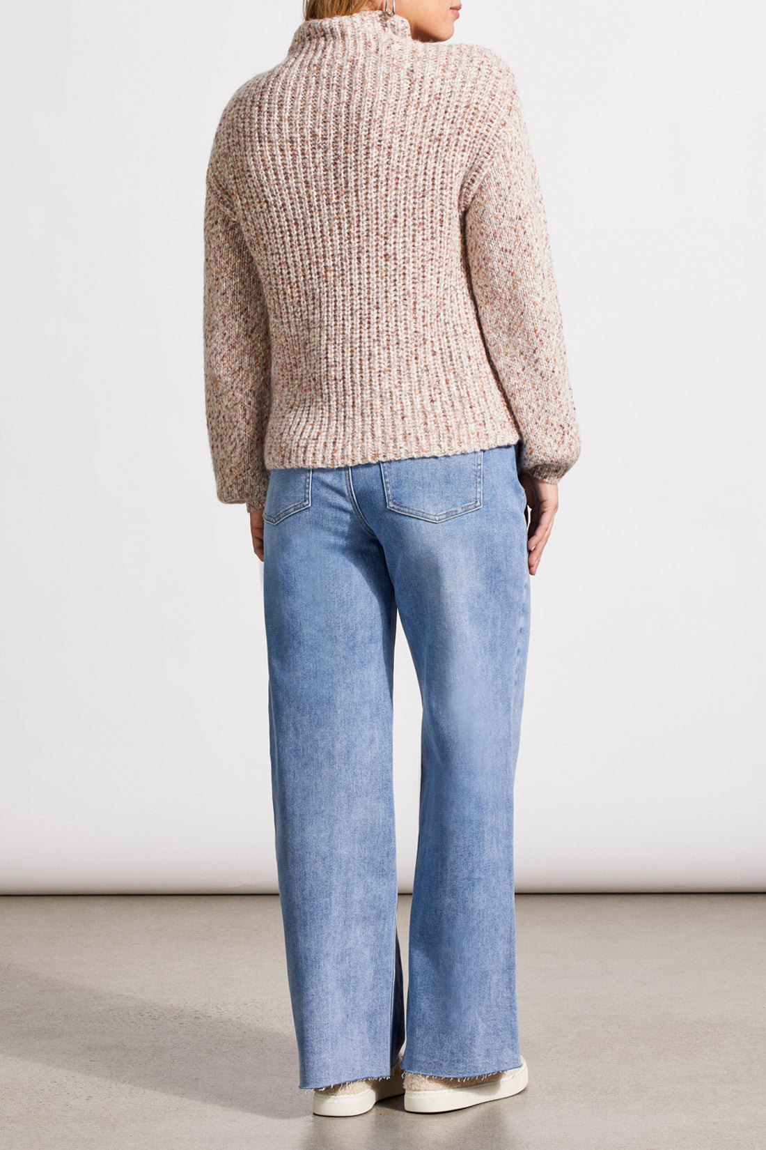 Person wearing a red patterned coat, cozy Tribal Funnel Neck Oversize Sweater in red, blue jeans, and black ankle boots stands against a dark paneled wall.