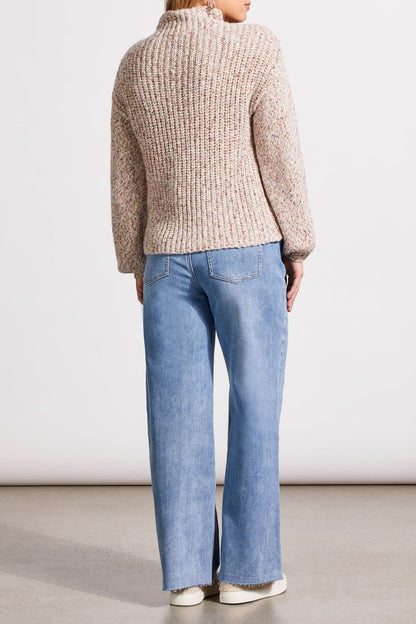 Person wearing a red patterned coat, cozy Tribal Funnel Neck Oversize Sweater in red, blue jeans, and black ankle boots stands against a dark paneled wall.
