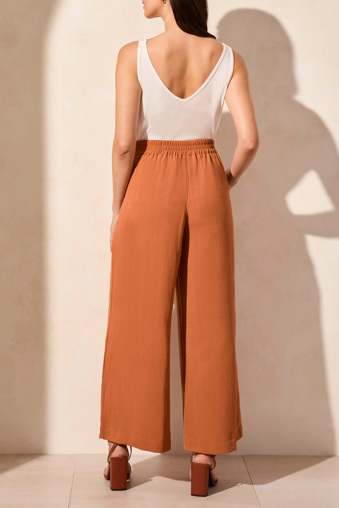 A woman stands against a neutral background wearing a rust-colored button-up shirt, a white top, brown heeled sandals, and Tribal’s FLOWY PANT WITH BLACK ELASTIC WAISTBAND.