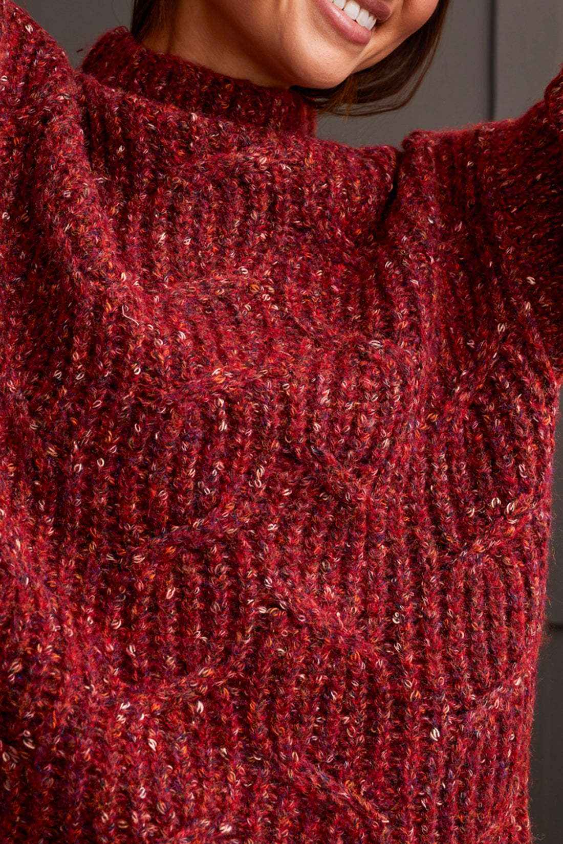 Person wearing a red patterned coat, cozy Tribal Funnel Neck Oversize Sweater in red, blue jeans, and black ankle boots stands against a dark paneled wall.