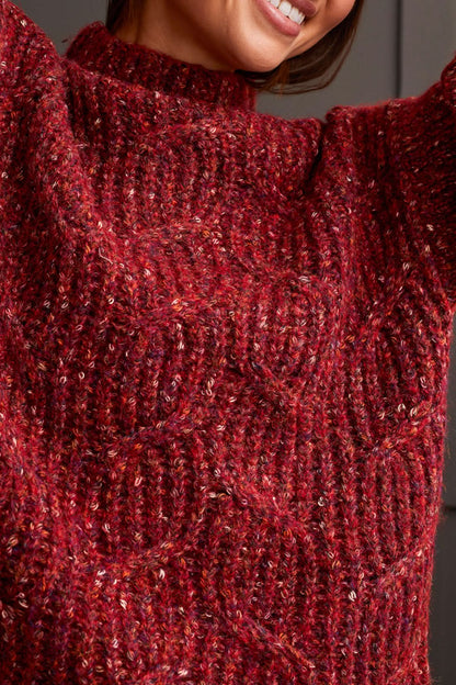 Person wearing a red patterned coat, cozy Tribal Funnel Neck Oversize Sweater in red, blue jeans, and black ankle boots stands against a dark paneled wall.
