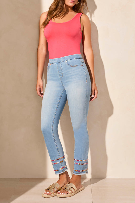 A woman stands against a beige wall wearing a sleeveless coral top, Tribal AUDREY PULL ON SLIM ANKLE JEANS with beaded hems for a flattering fit, and beige sandals—blending comfort and effortless style.