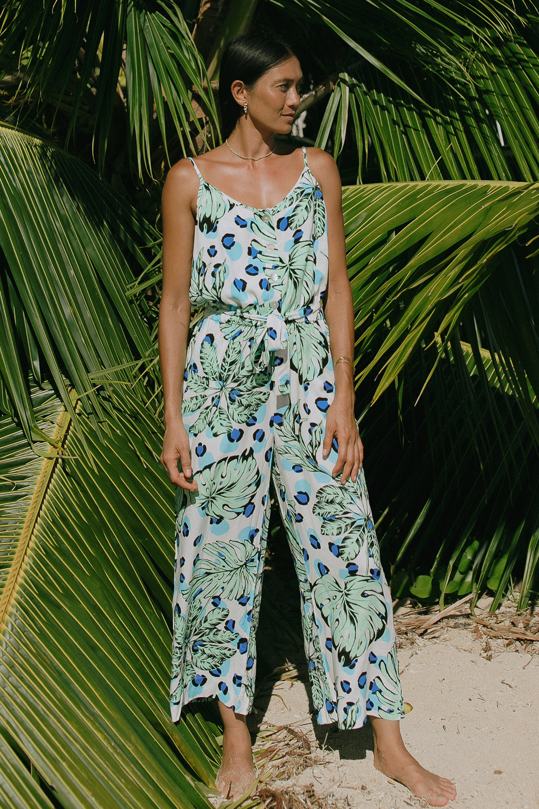 A woman stands barefoot on sand, wearing the Tribal BUTTON FRONT JUMPSUIT WITH POCKETS in a sleeveless tropical print—an effortless summer style surrounded by lush green palm leaves.