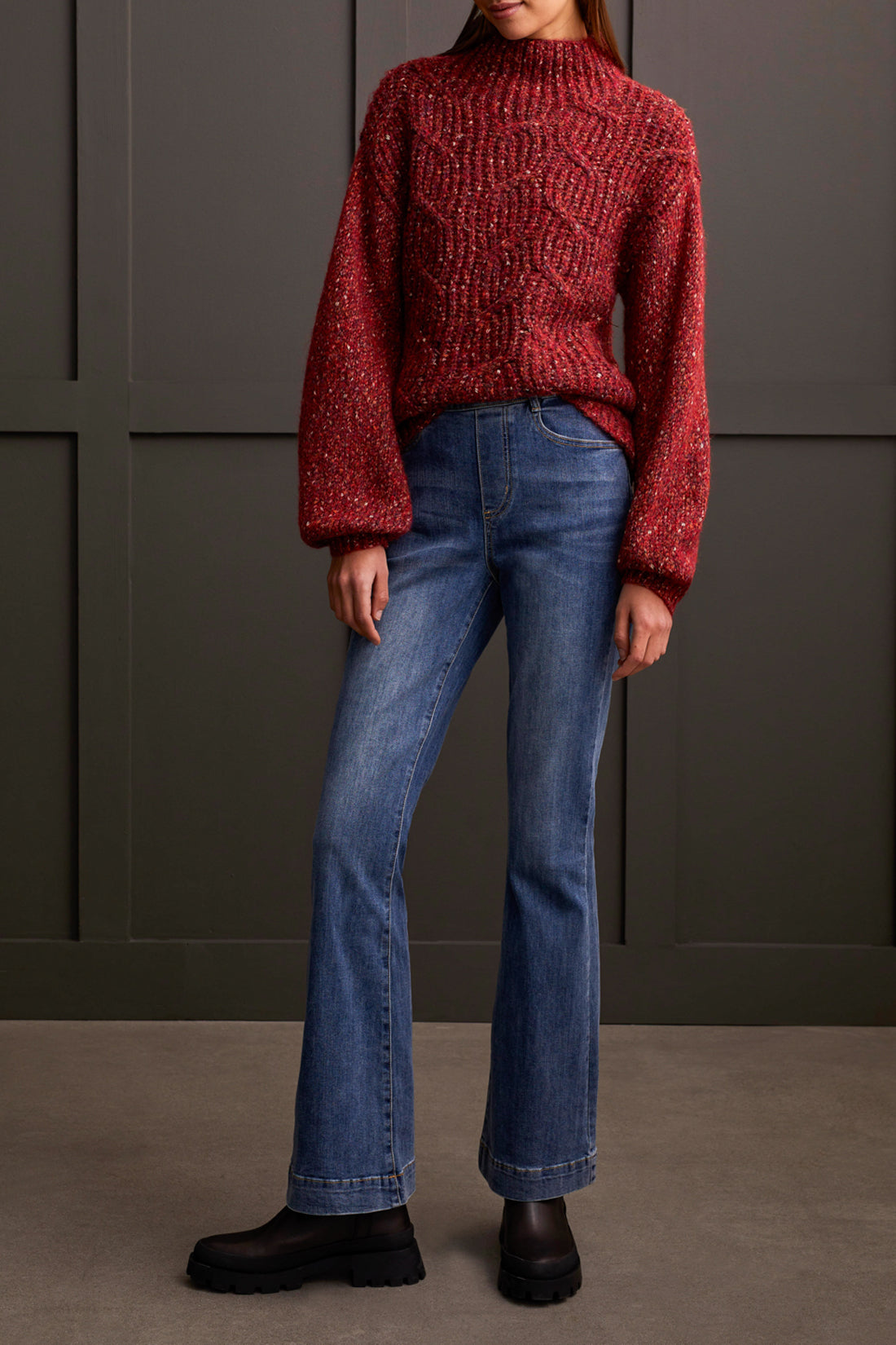 Person wearing a red patterned coat, cozy Tribal Funnel Neck Oversize Sweater in red, blue jeans, and black ankle boots stands against a dark paneled wall.