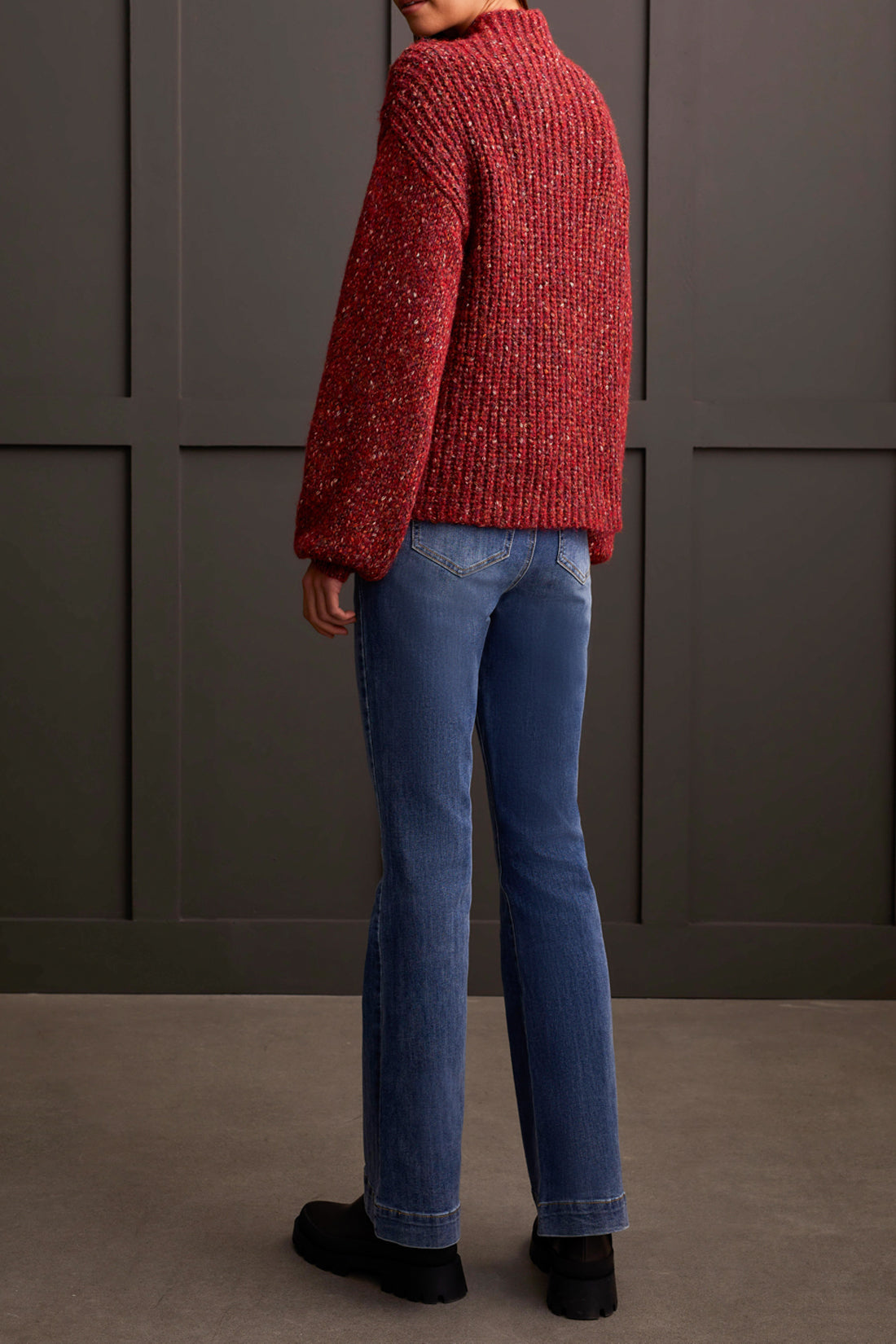 Person wearing a red patterned coat, cozy Tribal Funnel Neck Oversize Sweater in red, blue jeans, and black ankle boots stands against a dark paneled wall.