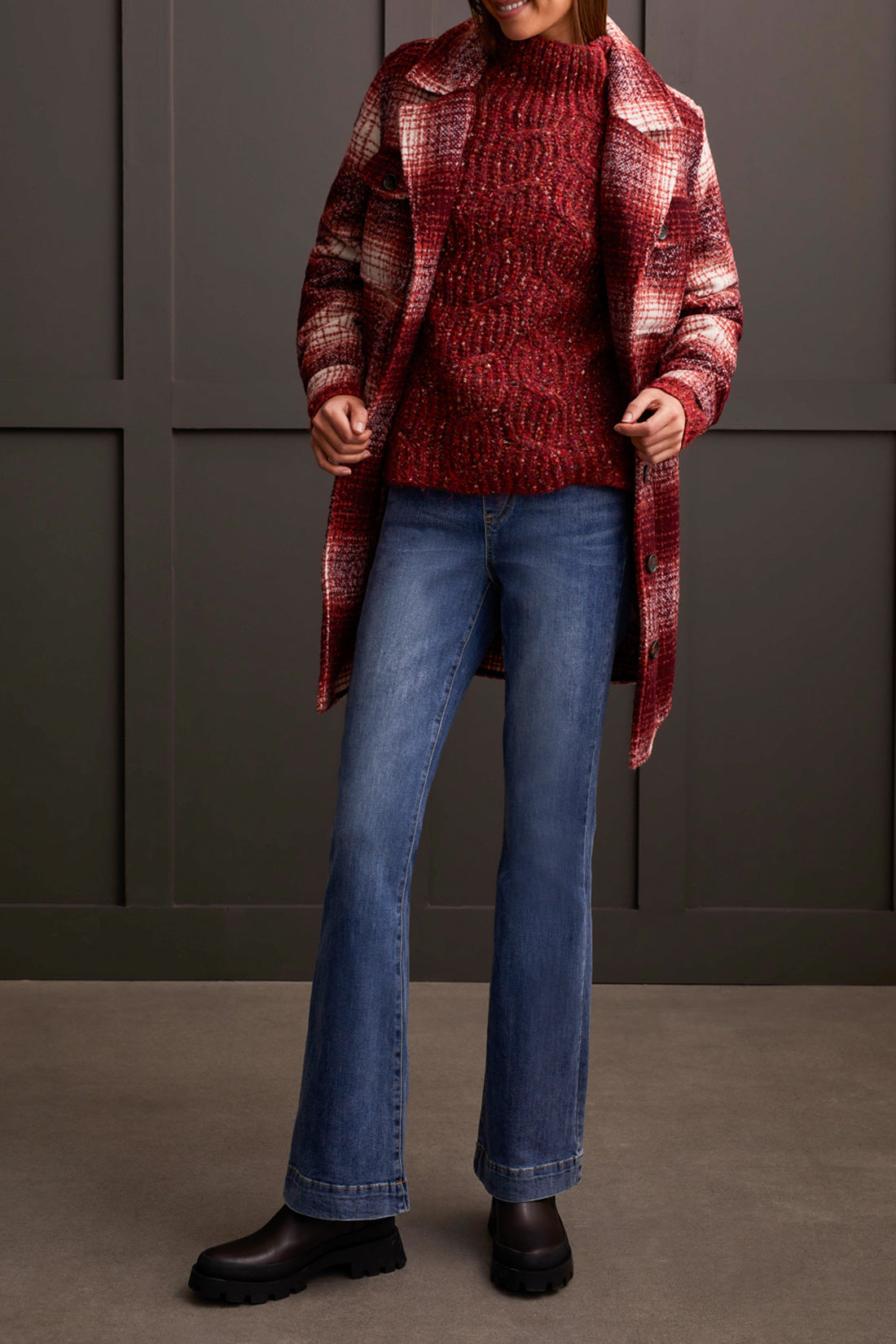 Person wearing a red patterned coat, cozy Tribal Funnel Neck Oversize Sweater in red, blue jeans, and black ankle boots stands against a dark paneled wall.