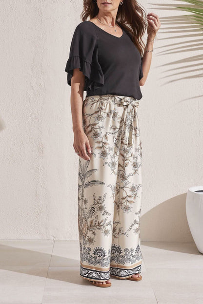 A woman stands outdoors, facing away, wearing a Tribal V Neck Raglan Combo Sleeved Top and patterned cream trousers with sandals.