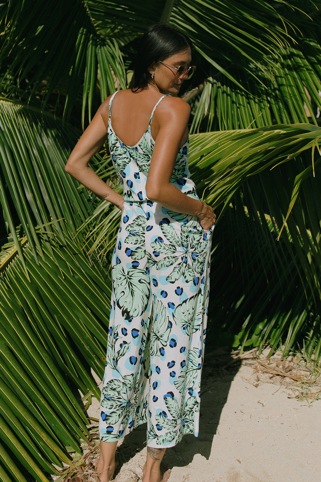 A woman stands barefoot on sand, wearing the Tribal BUTTON FRONT JUMPSUIT WITH POCKETS in a sleeveless tropical print—an effortless summer style surrounded by lush green palm leaves.