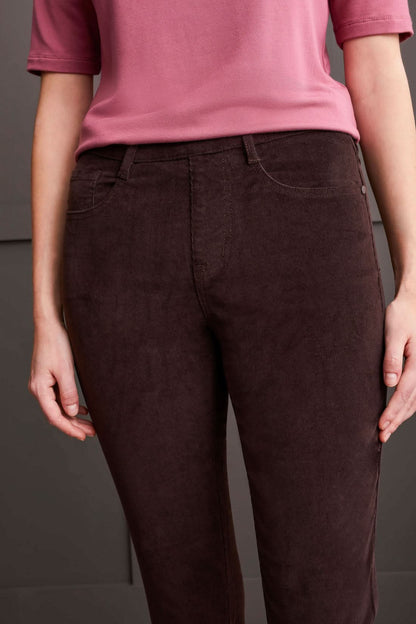 A person clad in a cream-colored quilted coat, a striped top, Tribal Audrey Pull on Slim Ankle Pants in burgundy, and white sneakers stands against a paneled grey wall.