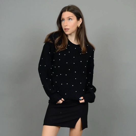 A person stands against a plain background wearing the stylish and cozy RD International Niri Long Sleeve Pull Over Sweater adorned with white dots, paired with a black skirt featuring a front slit. Their hands are in their pockets as they look slightly to their right.