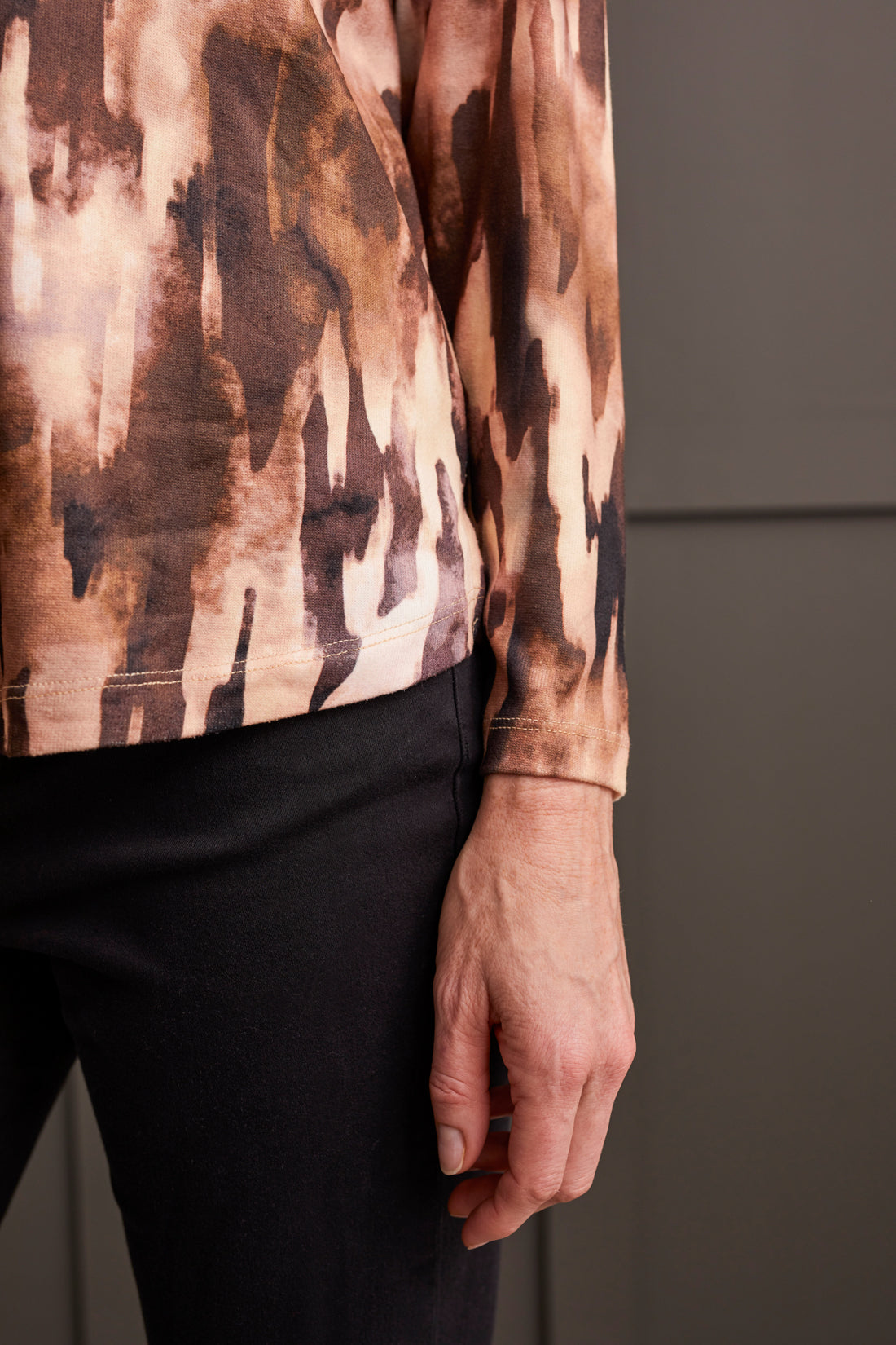A person wearing the Tribal Crew Neck Top with Curved Hem, featuring a stylish patterned design, paired with a black knee-length skirt and black ankle boots, stands against a dark paneled wall—perfect for any fall wardrobe.