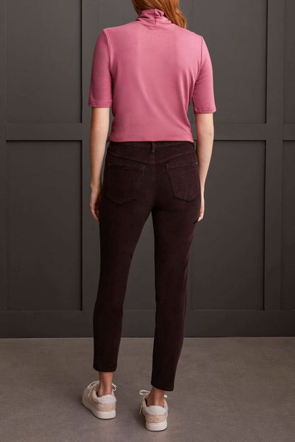 A person clad in a cream-colored quilted coat, a striped top, Tribal Audrey Pull on Slim Ankle Pants in burgundy, and white sneakers stands against a paneled grey wall.