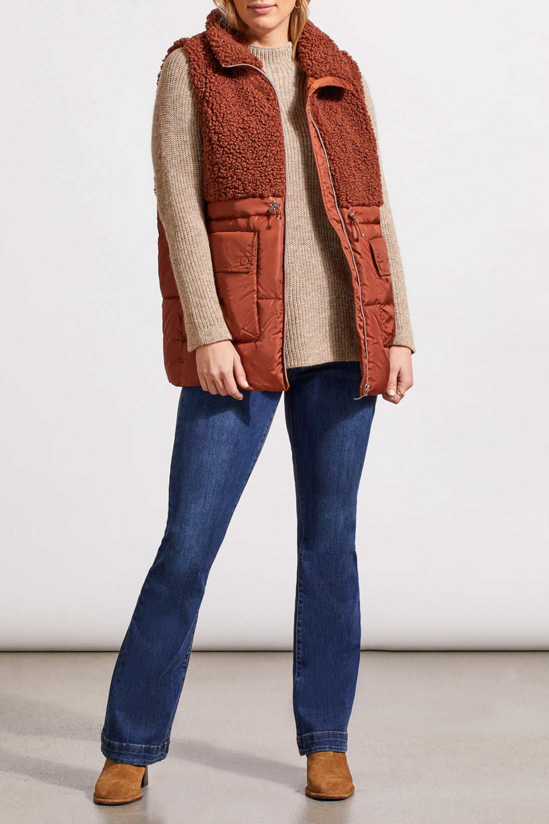 A person stands wearing a beige sweater, blue jeans, brown ankle boots, and a Tribal Sleeveless Sherpa Combo Nylon Vest in brown. The background is plain and white.