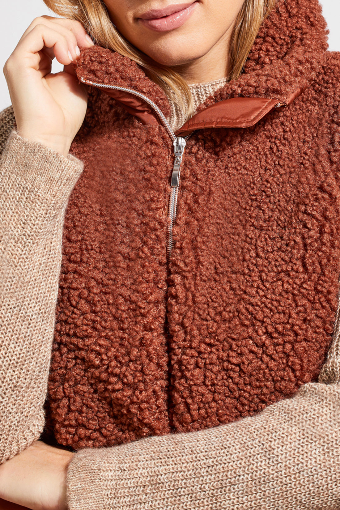 A person stands wearing a beige sweater, blue jeans, brown ankle boots, and a Tribal Sleeveless Sherpa Combo Nylon Vest in brown. The background is plain and white.
