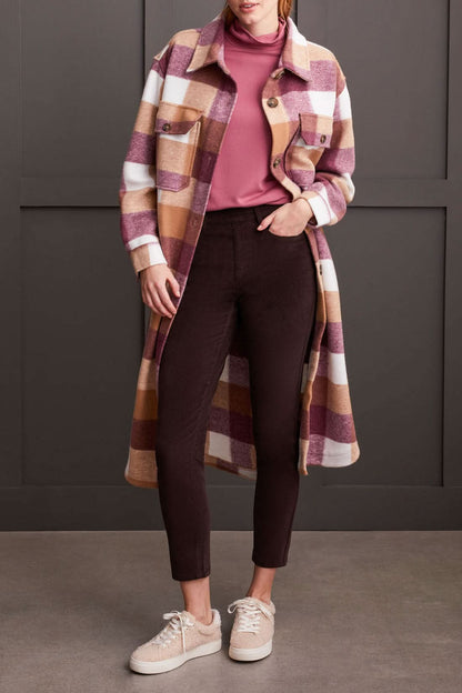 A person clad in a cream-colored quilted coat, a striped top, Tribal Audrey Pull on Slim Ankle Pants in burgundy, and white sneakers stands against a paneled grey wall.