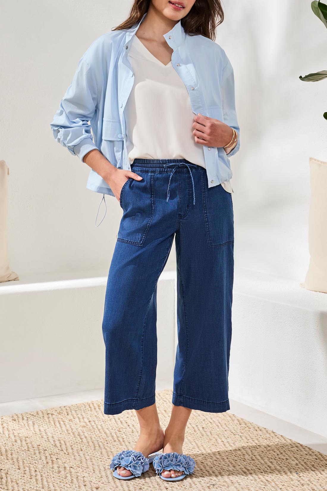 A woman stands indoors on a woven rug, wearing the Sophia Anke Jean by Tribal paired with a light blue jacket, white top, and ruffle-detailed sandals.