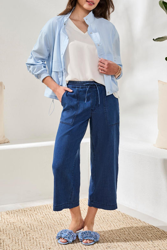 A woman stands indoors on a woven rug, wearing the Sophia Anke Jean by Tribal paired with a light blue jacket, white top, and ruffle-detailed sandals.