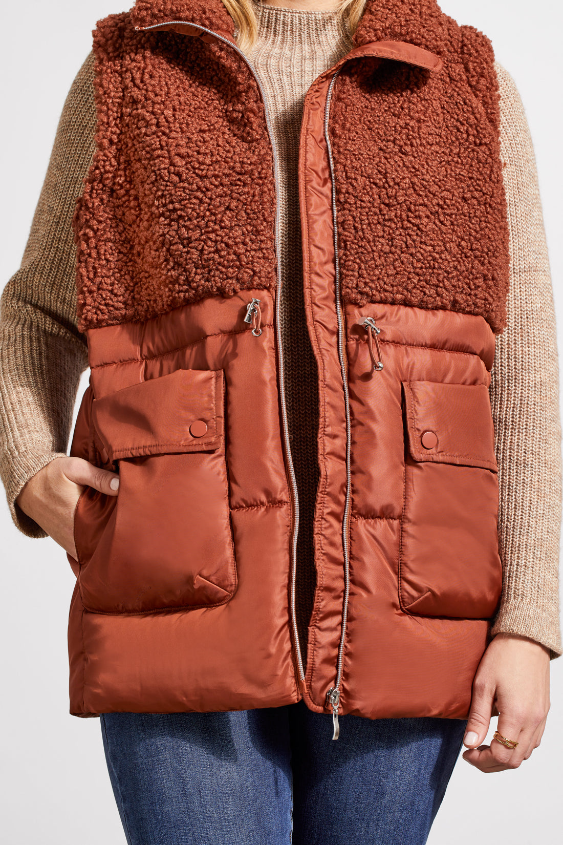 A person stands wearing a beige sweater, blue jeans, brown ankle boots, and a Tribal Sleeveless Sherpa Combo Nylon Vest in brown. The background is plain and white.