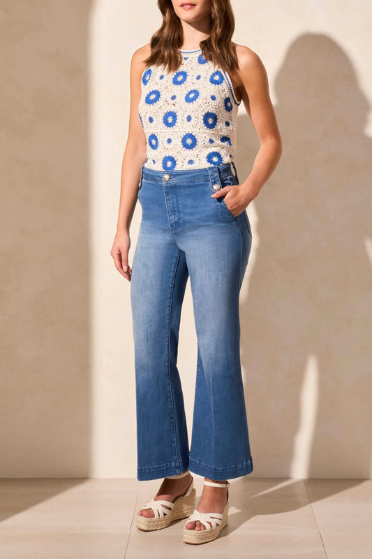 A person wearing a white and blue floral sleeveless top, Tribal’s BROOKE HUGGING PALAZZO MARINE JEANS, and white wedge sandals stands against a light-colored wall.