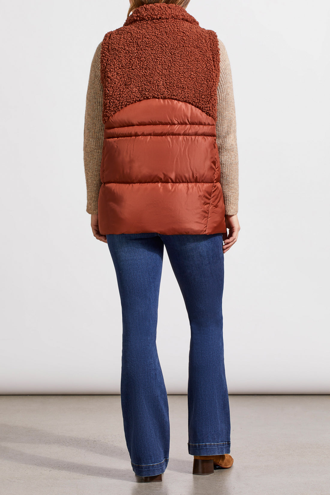 A person stands wearing a beige sweater, blue jeans, brown ankle boots, and a Tribal Sleeveless Sherpa Combo Nylon Vest in brown. The background is plain and white.