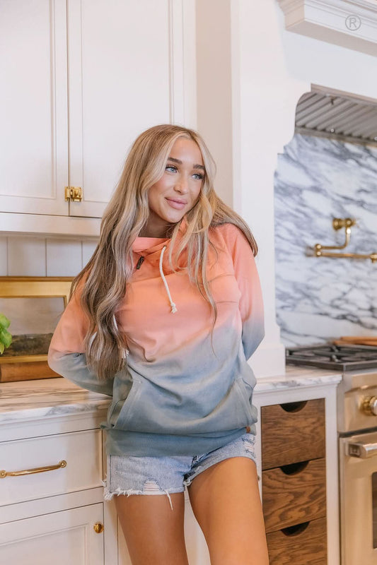 Dressed in Ampersand Avenue's Chasing Sunset Doublehooded Sweatshirt and denim shorts, a person exudes comfort and style while standing in a kitchen adorned with a marble backsplash and wooden cabinets.