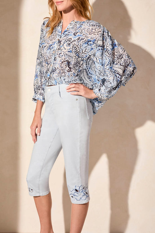 Woman wearing a blue patterned blouse and Tribal SOPHIA CURVY CAPRI JEANS—stylish capris with embroidered cuffs—paired with beige heeled sandals, standing against a plain light background.