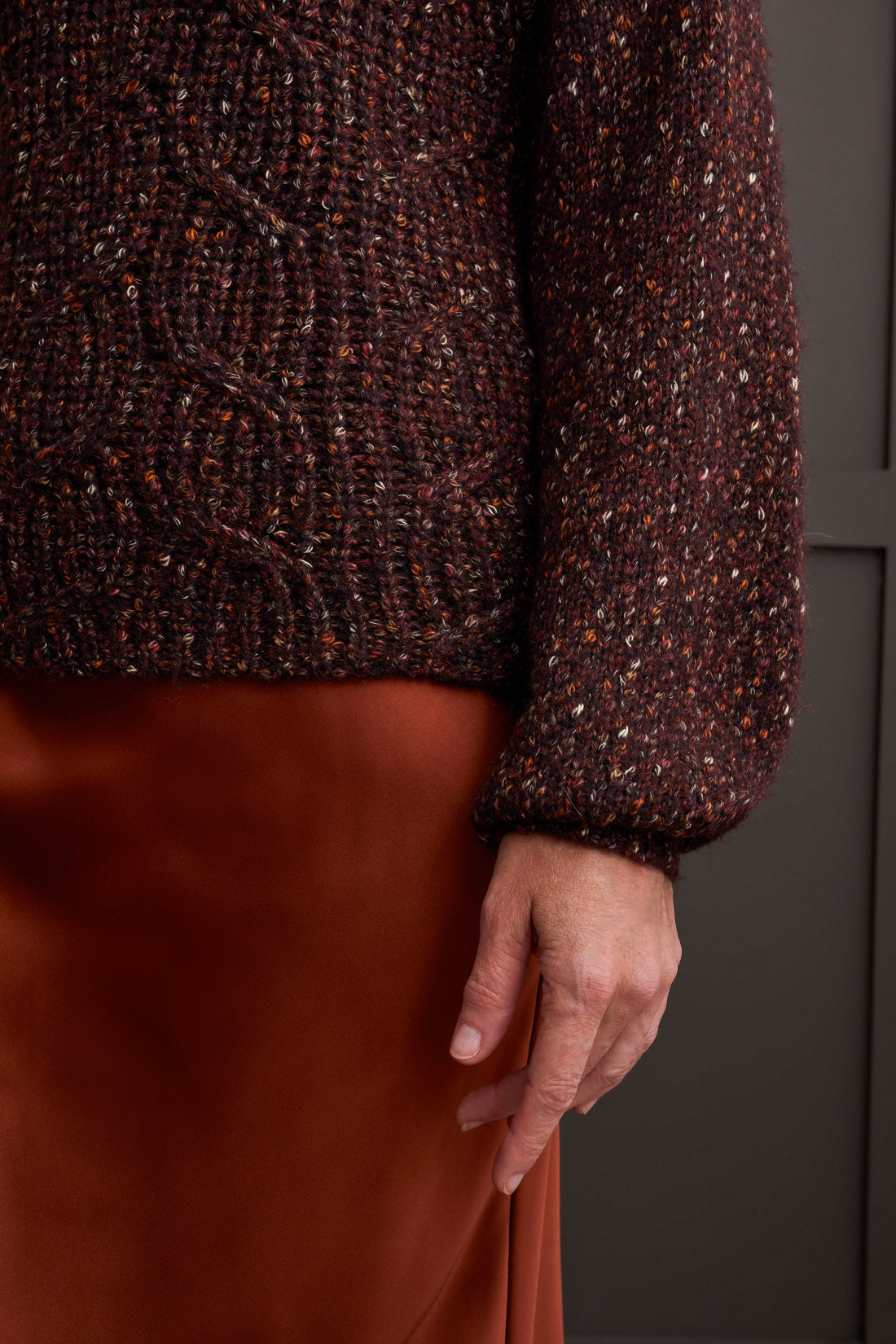 Person wearing a red patterned coat, cozy Tribal Funnel Neck Oversize Sweater in red, blue jeans, and black ankle boots stands against a dark paneled wall.